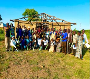 a big group of adults stand in front of a wood shack facing camera
