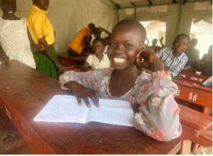 girl sitting at desk smiling widely at the camera, there is a notepad on the desk in front of her. She is wearing a grey and orange top