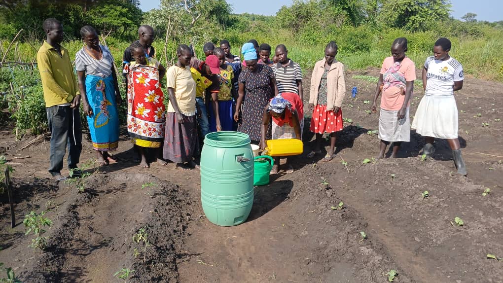 large group standing in field around a green water butt.