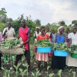 Group is in a field facing camera. They are holding full baskets of green produce.