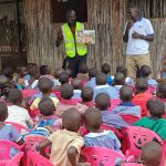 man in yellow high vis jacket and man in grey t-shirt teaching group of children in multi-coloured uniform. Children are sat in a group facing away from camera