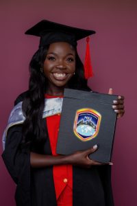 burgundy background. Woman smiles at camera wearing black graduation cap with red tassel and gown and red dress. She presents her diploma to camera