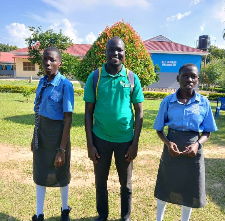 man in green top smiling. standing between two students in blue polo tops and long grey skirts
