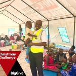 man in yellow high-vis jacket, holding toddler. they are in a clinic marquee. Patients lay around them. The Big Give xmas logo is in bottom left corner