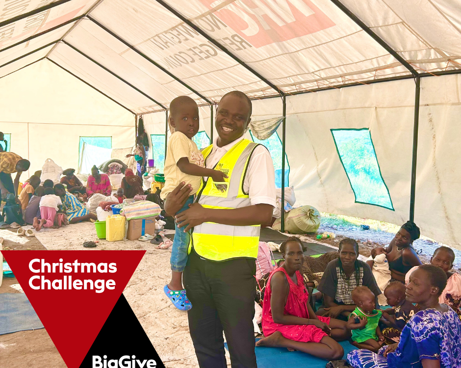 man in yellow high-vis jacket, holding toddler. they are in a clinic marquee. Patients lay around them. The Big Give xmas logo is in bottom left corner