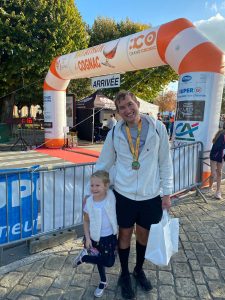 man and daughter stand next to a orange and white finishing line. Man is wearing white and black running gear with a medal on his chest. Girl is also in black and white sports gear.