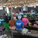 Group sat facing a whiteboard at the back of a room. The room has grey walls and a plastic roof with wooden beams. They are sat on built in benches.