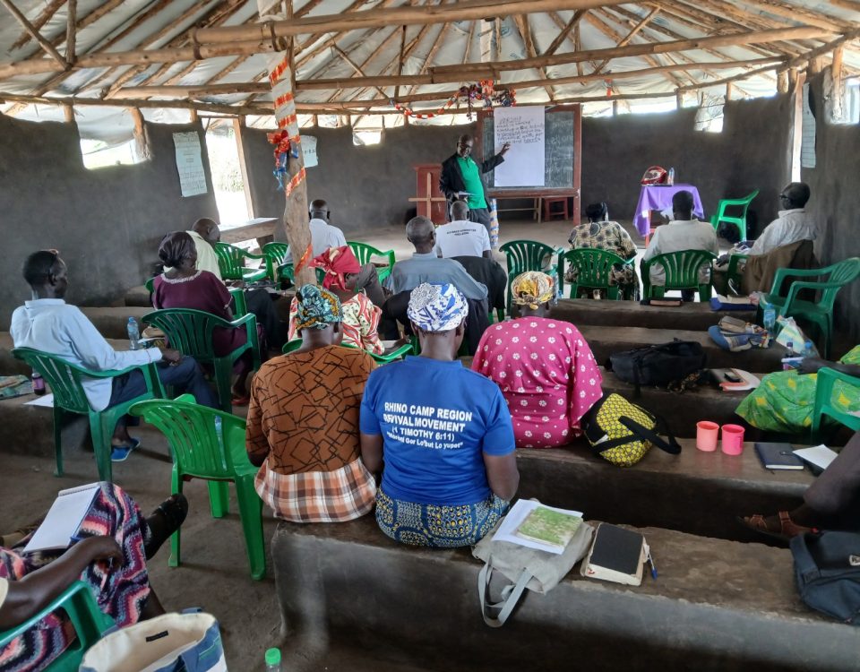Group sat facing a whiteboard at the back of a room. The room has grey walls and a plastic roof with wooden beams. They are sat on built in benches.