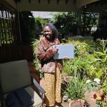 Smiling woman in leopard print coat and yellow/green patterned trousers standing in a sunny courtyard filled with plants. She is holding a laptop in one hand and doing a thumbs-up with other hand.