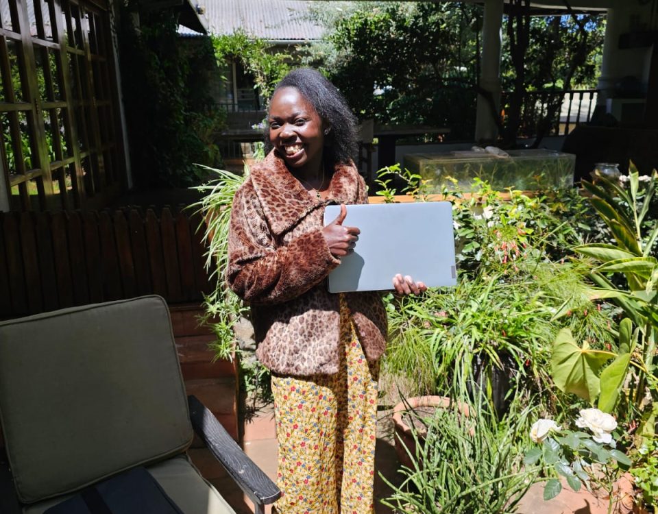 Smiling woman in leopard print coat and yellow/green patterned trousers standing in a sunny courtyard filled with plants. She is holding a laptop in one hand and doing a thumbs-up with other hand.