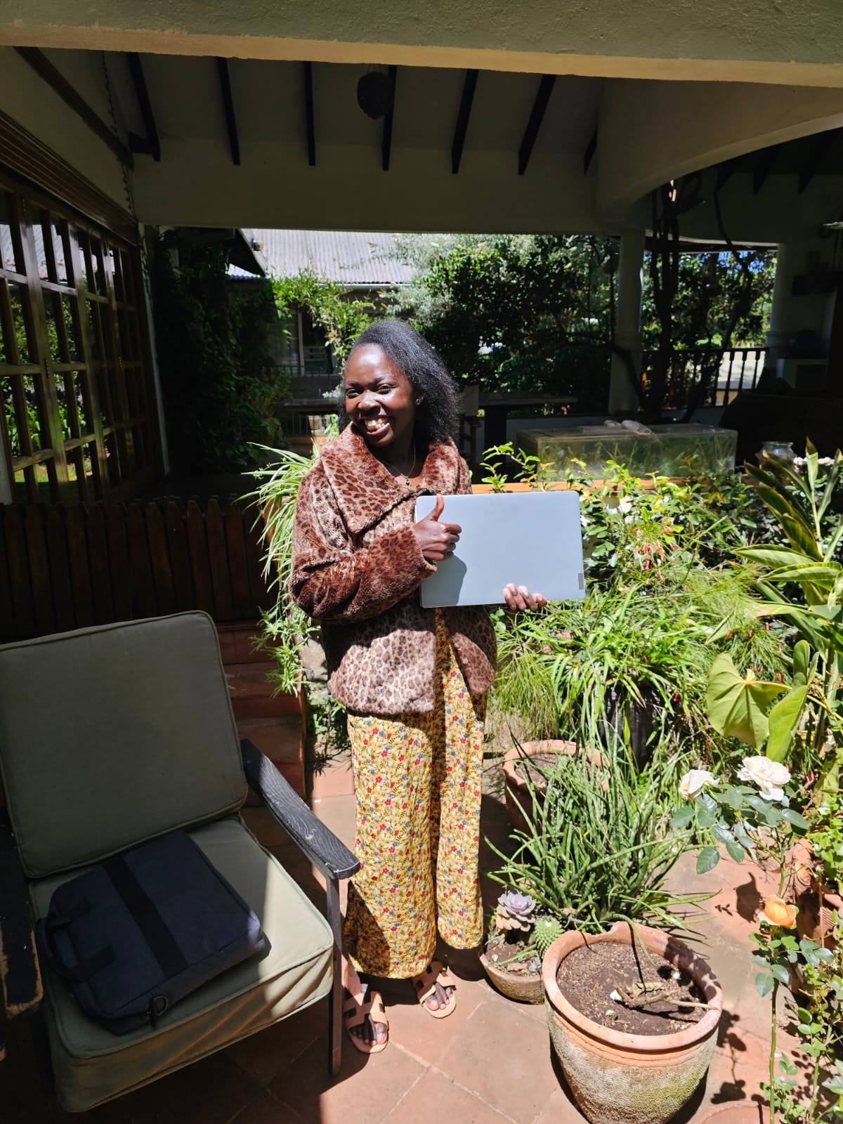 Smiling woman in leopard print coat and yellow/green patterned trousers standing in a sunny courtyard filled with plants. She is holding a laptop in one hand and doing a thumbs-up with other hand.