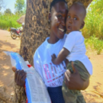 woman standing under tree in shade. She is wearing a white graphic print t-shirt and blue bottoms. In one arm, she is holding her baby in a white onsie. Their faces are pressed together. In the other hand she has a plastic kit bag with blue edging.