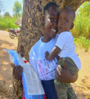 woman standing under tree in shade. She is wearing a white graphic print t-shirt and blue bottoms. In one arm, she is holding her baby in a white onsie. Their faces are pressed together. In the other hand she has a plastic kit bag with blue edging.