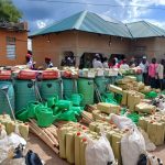 water butts, watering cans, tools, fertilizers, seeds piled before a yellow building with a green roof.