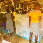 Mothers demonstrating how to hang mosquito net in building with grass roof. They are presenting to a group sat at the back of the space. The mosquito net is hung in front of them