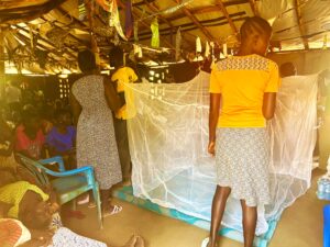 Mothers demonstrating how to hang mosquito net in building with grass roof. They are presenting to a group sat at the back of the space. The mosquito net is hung in front of them