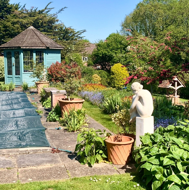 picture of a established garden. with sculpture of a woman, a covered pool, and a teal garden house at the end of the pool