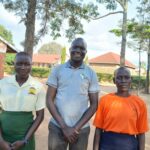 man stands between two students. the man is wearing a grey polo shirt with CRESS logo and brown trousers. On left, student is wearing a yellow shirt with a green skirt. On the rights, the student is wearing a bright orange t-shirt and navy skirt