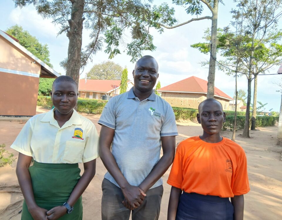 man stands between two students. the man is wearing a grey polo shirt with CRESS logo and brown trousers. On left, student is wearing a yellow shirt with a green skirt. On the rights, the student is wearing a bright orange t-shirt and navy skirt