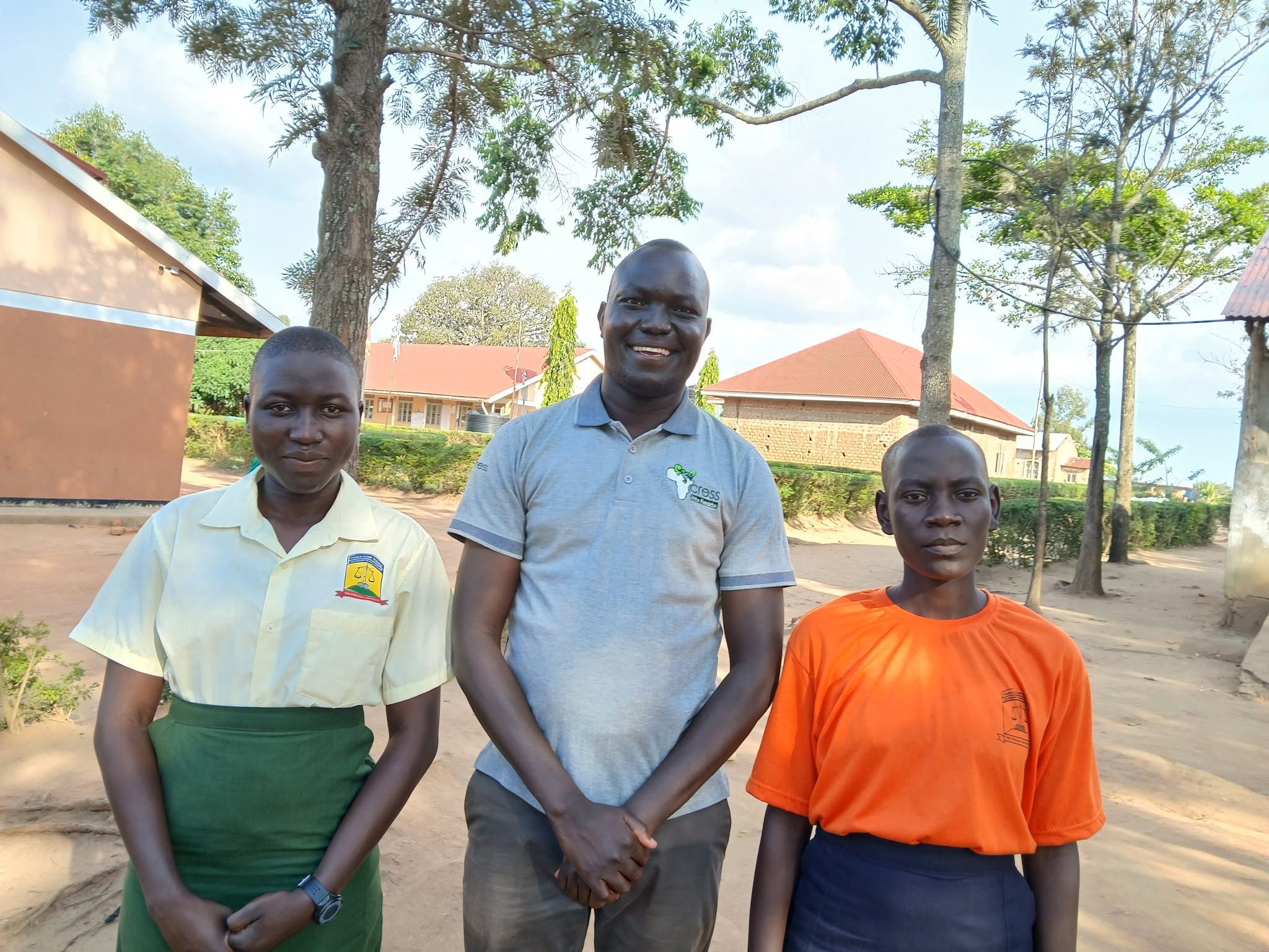 man stands between two students. the man is wearing a grey polo shirt with CRESS logo and brown trousers. On left, student is wearing a yellow shirt with a green skirt. On the rights, the student is wearing a bright orange t-shirt and navy skirt