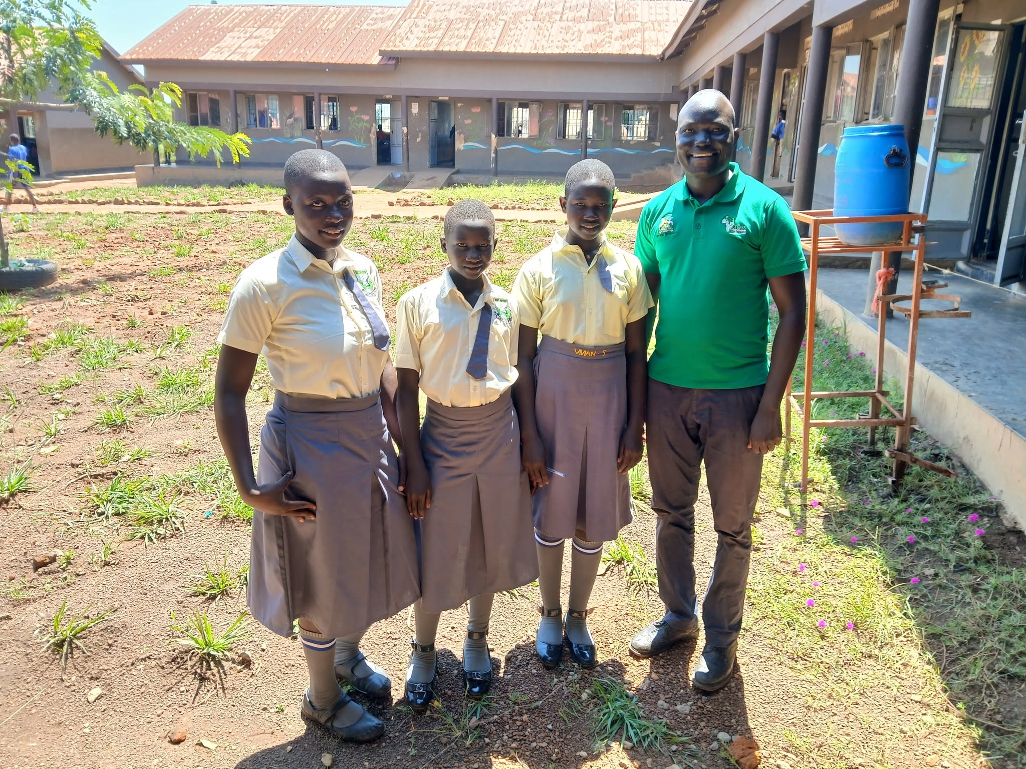 man stands with a group of students. students are wearing butter yellow shirts with brown skirts, socks and black shoes. Man is wearing green polo with logo and brown trousers