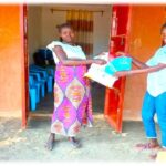 two women stand outside a red clay building. one on left wearing white top with pink patterned skirt. other on right wearing jeans and white shirt. woman on right is handing package to other woman.