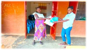 two women stand outside a red clay building. one on left wearing white top with pink patterned skirt. other on right wearing jeans and white shirt. woman on right is handing package to other woman.