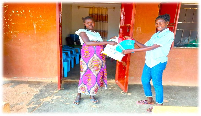 two women stand outside a red clay building. one on left wearing white top with pink patterned skirt. other on right wearing jeans and white shirt. woman on right is handing package to other woman.