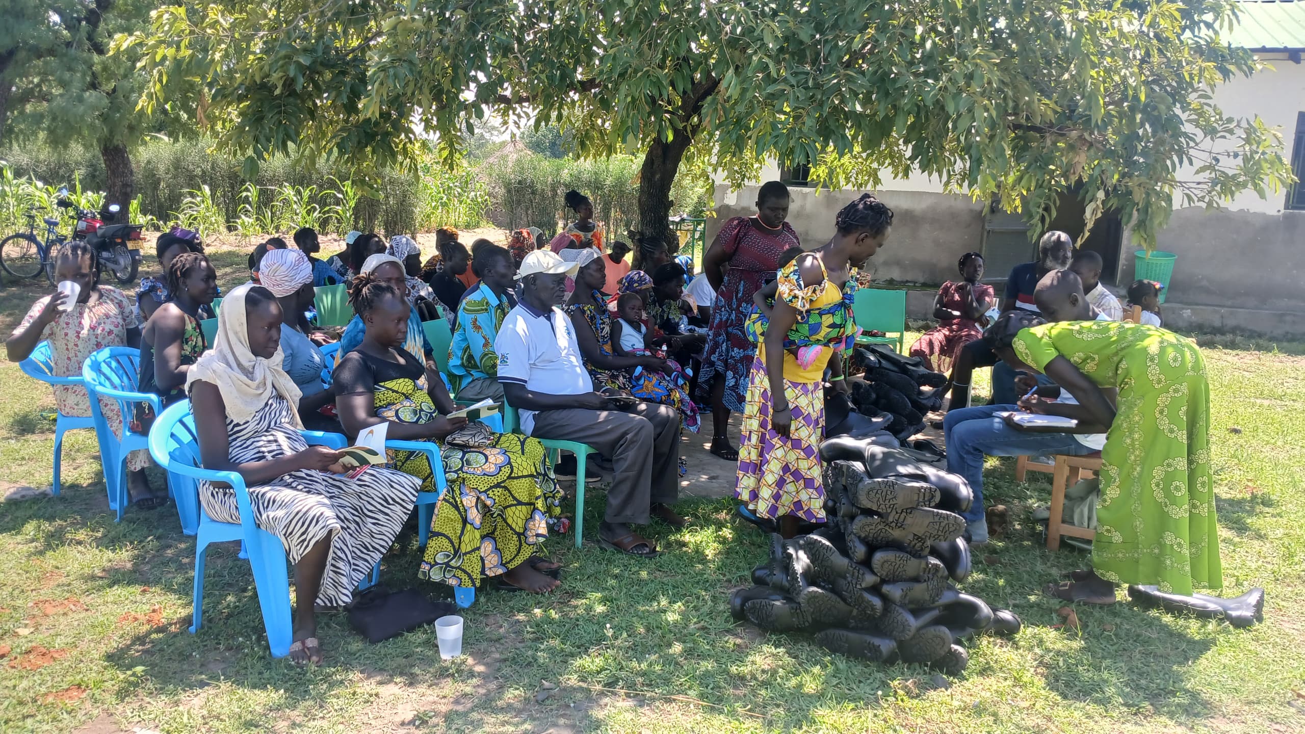 group sat on chairs arranged under a tree waiting to be given new agricultural equipment, including gum boots