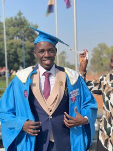 man smiling at camera, adjusting robes. He is wearing a bright blue graduation cap and gown with beige accents. He is also wearing a navy suit with a white shirt and red patterned tie. Someone off camera is holding the cap tassle up