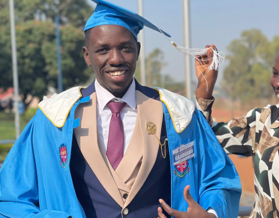 man smiling at camera, adjusting robes. He is wearing a bright blue graduation cap and gown with beige accents. He is also wearing a navy suit with a white shirt and red patterned tie. Someone off camera is holding the cap tassle up