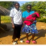 two women standing next to a grass roofed home. One is wearing a white polo with green collar, black trousers and white shoes. The other woman has a bright blue top with a navy patterned skirt and red shoes. She is holding a baby in a red blanket