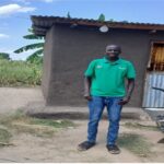 Man standing in front of single story house with a tin roof. He is wearing blue jeans and a green shirt with logo. next to him is a yellow and red motorbike