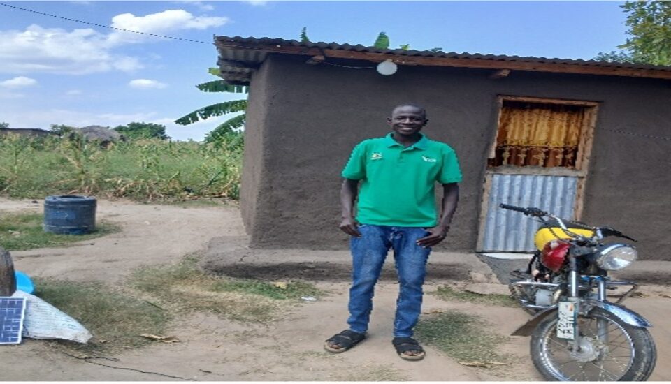 Man standing in front of single story house with a tin roof. He is wearing blue jeans and a green shirt with logo. next to him is a yellow and red motorbike
