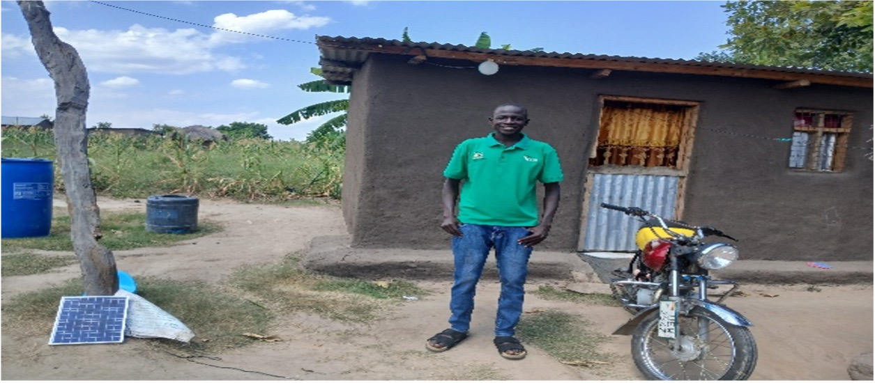 Man standing in front of single story house with a tin roof. He is wearing blue jeans and a green shirt with logo. next to him is a yellow and red motorbike
