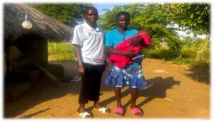 two women standing next to a grass roofed home. One is wearing a white polo with green collar, black trousers and white shoes. The other woman has a bright blue top with a navy patterned skirt and red shoes. She is holding a baby in a red blanket