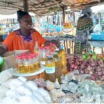 woman in orange shirt with purple collar standing behind a market stall table. There are bottles of cooking oil, onions, tomatoes, ginger, and small bags of flour, beans, and grains. There is a wide green bucket with flour in that she is measuring out.