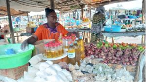 woman in orange shirt with purple collar standing behind a market stall table. There are bottles of cooking oil, onions, tomatoes, ginger, and small bags of flour, beans, and grains. There is a wide green bucket with flour in that she is measuring out.