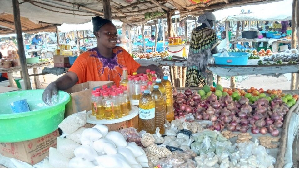 woman in orange shirt with purple collar standing behind a market stall table. There are bottles of cooking oil, onions, tomatoes, ginger, and small bags of flour, beans, and grains. There is a wide green bucket with flour in that she is measuring out.