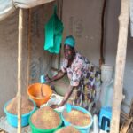 woman in market stall. Canvas rolled up above her. Green bags hanging next to her. There are large green and blue bucket of grain on the table in front of her with blue cups for measuring. She is wearing a turquoise head wrap, a pink floral top and a red, yellow and black patterned skirt