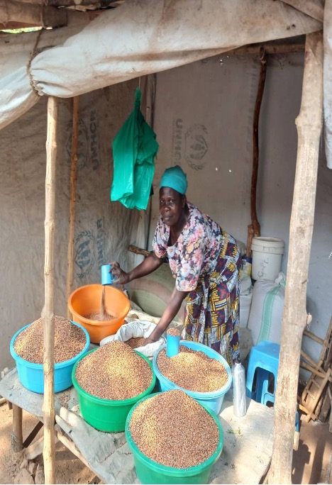 woman in market stall. Canvas rolled up above her. Green bags hanging next to her. There are large green and blue bucket of grain on the table in front of her with blue cups for measuring. She is wearing a turquoise head wrap, a pink floral top and a red, yellow and black patterned skirt