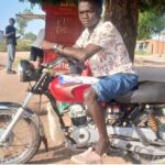 man sat on red motorbike with brown leather seat. it is parked under a tree next to a food stall. the man is wearing a white shirt with red fauna pattern, light blue denim shorts and pink trainers