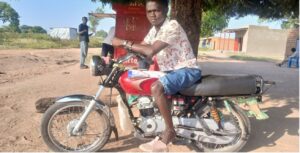 man sat on red motorbike with brown leather seat. it is parked under a tree next to a food stall. the man is wearing a white shirt with red fauna pattern, light blue denim shorts and pink trainers