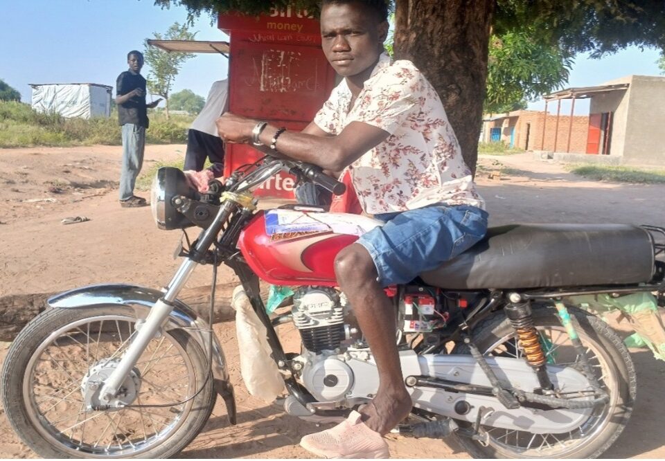 man sat on red motorbike with brown leather seat. it is parked under a tree next to a food stall. the man is wearing a white shirt with red fauna pattern, light blue denim shorts and pink trainers
