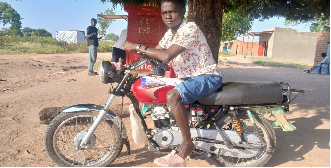 man sat on red motorbike with brown leather seat. it is parked under a tree next to a food stall. the man is wearing a white shirt with red fauna pattern, light blue denim shorts and pink trainers