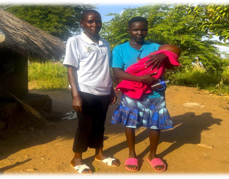 two women standing next to a grass roofed home. One is wearing a white polo with green collar, black trousers and white shoes. The other woman has a bright blue top with a navy patterned skirt and red shoes. She is holding a baby in a red blanket