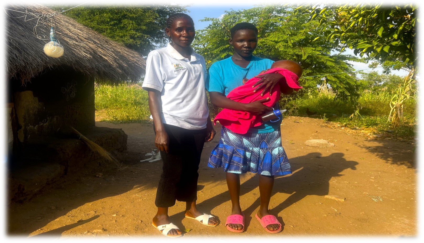 two women standing next to a grass roofed home. One is wearing a white polo with green collar, black trousers and white shoes. The other woman has a bright blue top with a navy patterned skirt and red shoes. She is holding a baby in a red blanket