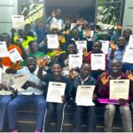 a group of 20 sitting on steps outside green building. They are holding certificates and smiling