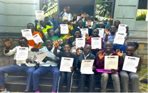 a group of 20 sitting on steps outside green building. They are holding certificates and smiling