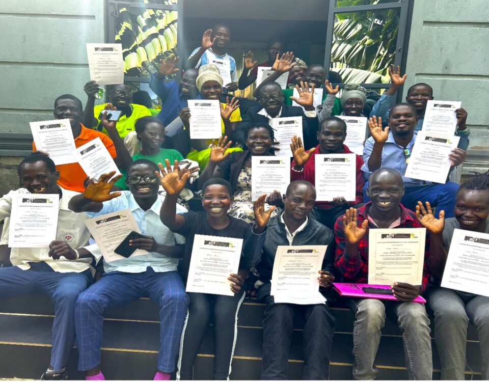 a group of 20 sitting on steps outside green building. They are holding certificates and smiling